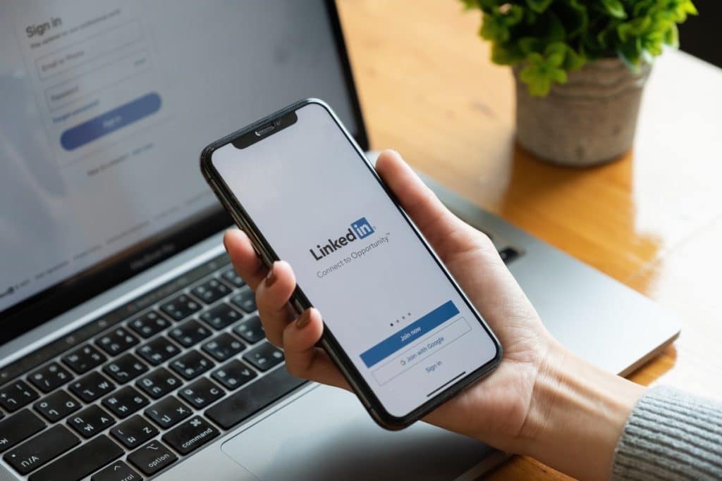 Elinde LinkedIn uygulaması açık akıllı telefon tutan kişi ve dizüstü bilgisayar – LinkedIn profili nasıl olmalı
